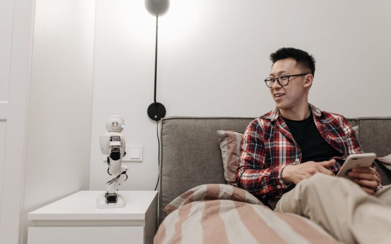 Man sitting on bed using mobile phone, interacting with small robot on bedside table.