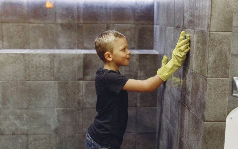 Side view of kid with blond hair in yellow rubber gloves cleaning tile wall in bathroom