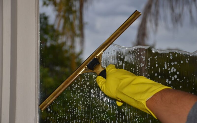 Close-up of hand cleaning window with squeegee outdoors using yellow gloves.