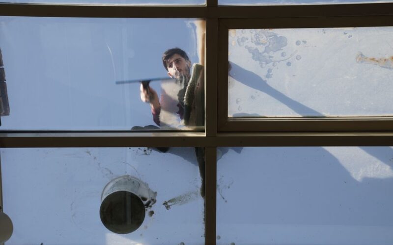 A male window cleaner washing the exterior glass roof from above with a tool during daylight.