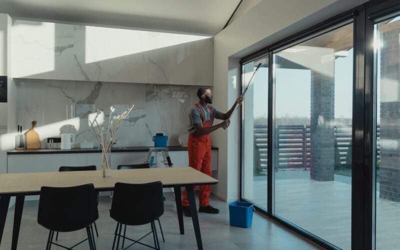 A professional cleaner in uniform washing large glass windows indoors with natural light streaming in.