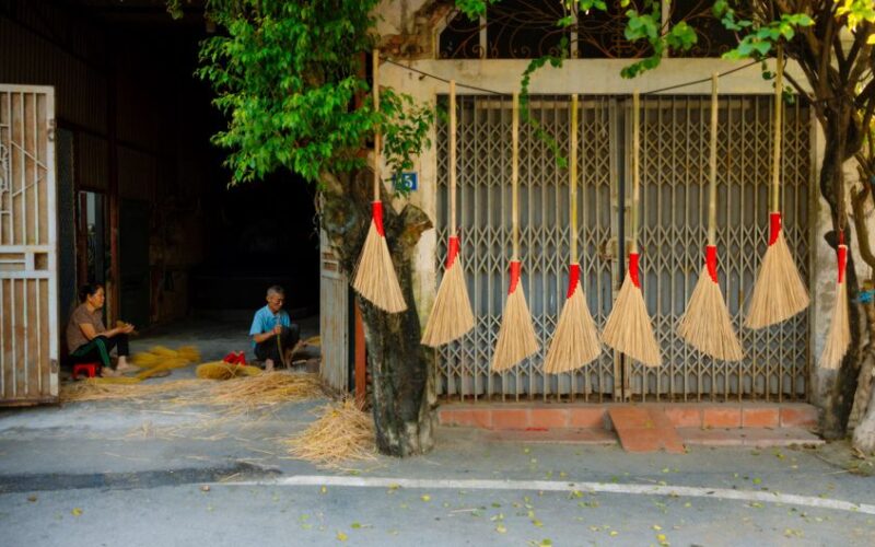 Two people making brooms outside a rural workshop with brooms hanging on the wall.