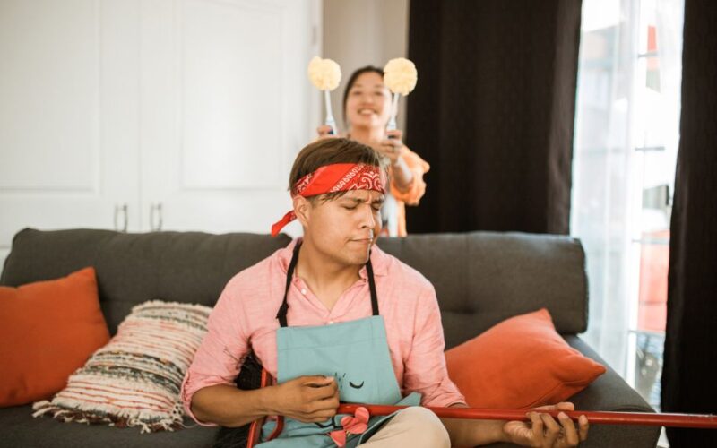 A playful scene of a man pretending to play a broom guitar while cleaning, with another person in the background.