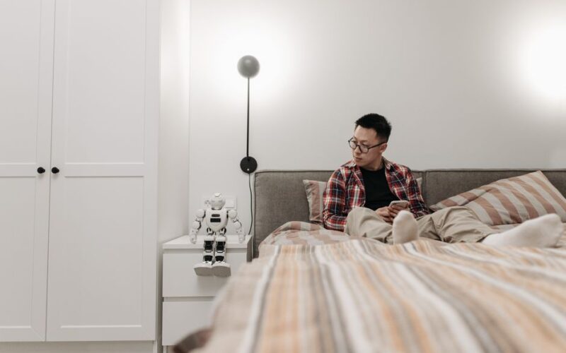 A man in a modern bedroom sitting on the bed with a robot toy beside him, creating a tech-savvy atmosphere.