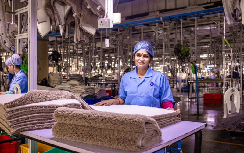 A female worker in a textile factory smiling, showcasing a stack of carpets.