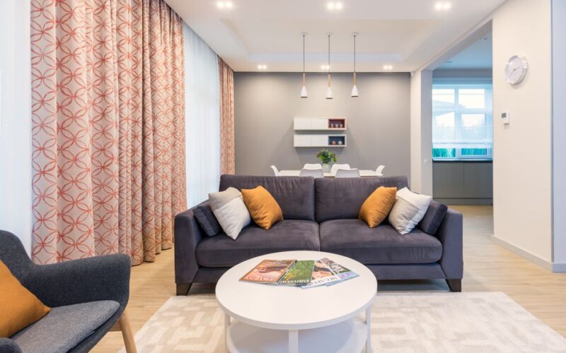 Comfortable sofa and armchair placed on carpet near side table with magazines and kitchen table in stylish spacious apartment with window
