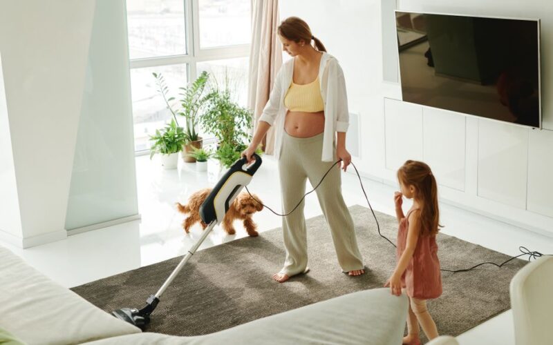 Pregnant mother cleaning carpet with vacuum as daughter and dog play indoors.