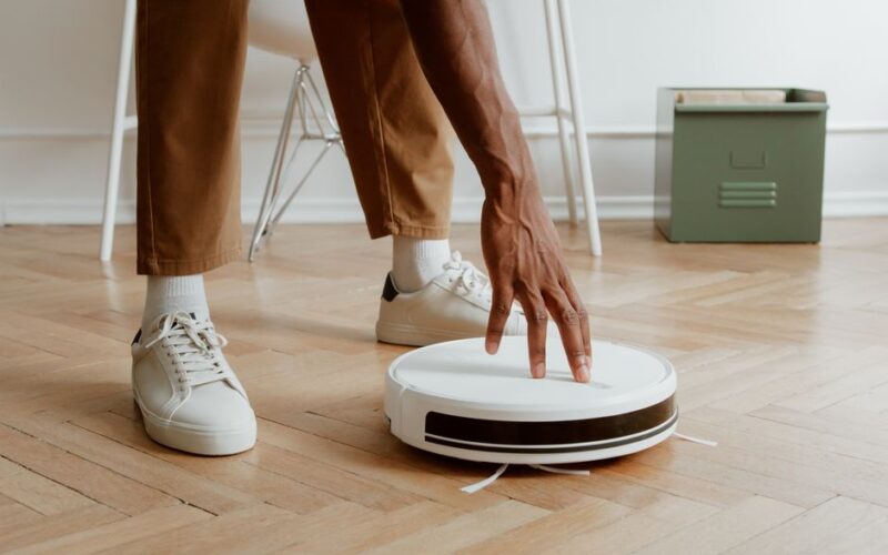A man's hand adjusting a robot vacuum on a wooden floor in a modern home interior.