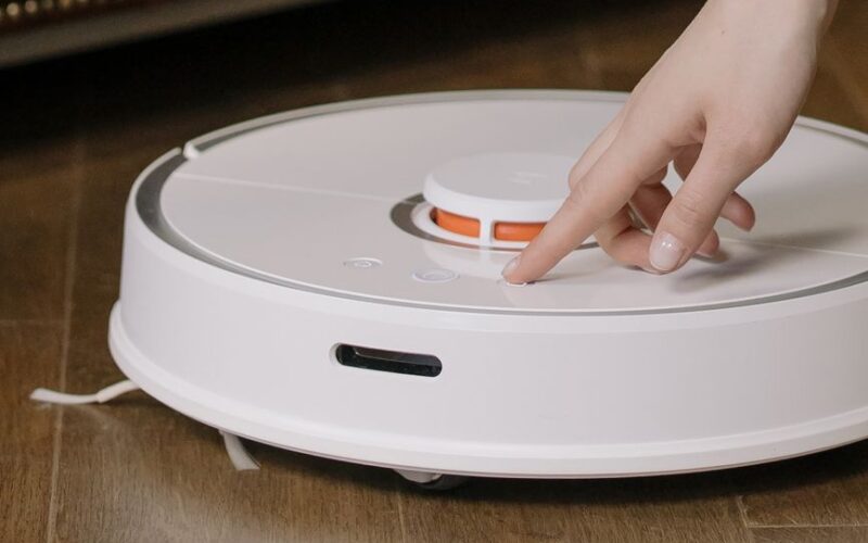 Close-up of a hand pressing a button on a robotic vacuum cleaner on a wooden floor.