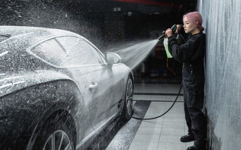 An Asian woman using a pressure washer to clean a car at an indoor car wash facility.