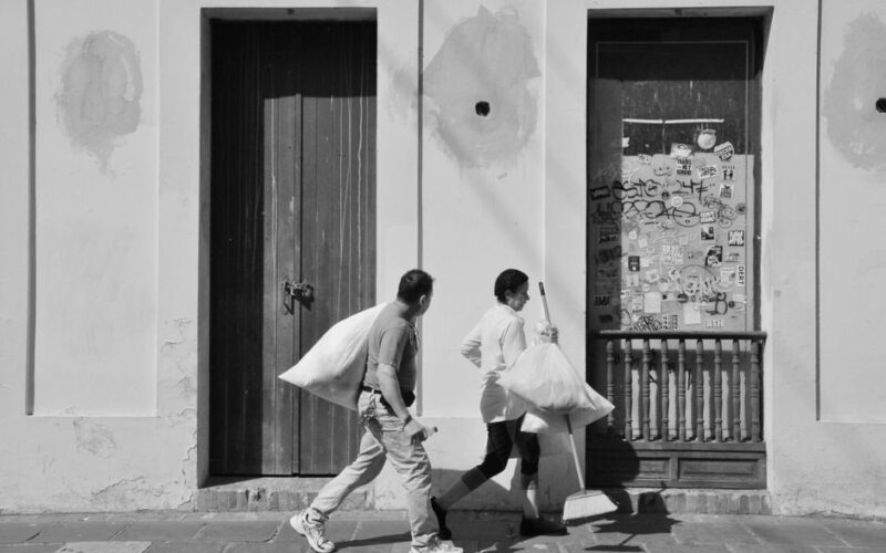 Two street cleaning workers carry bags past a graffiti-covered wall in black and white.