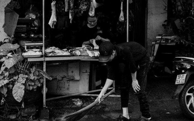 Black and white photo of a market vendor cleaning outside a shop.