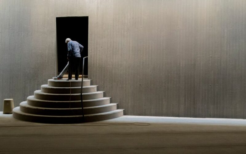 A solitary man cleaning stairs in a minimalist concrete interior, creating a sense of solitude and symmetry.