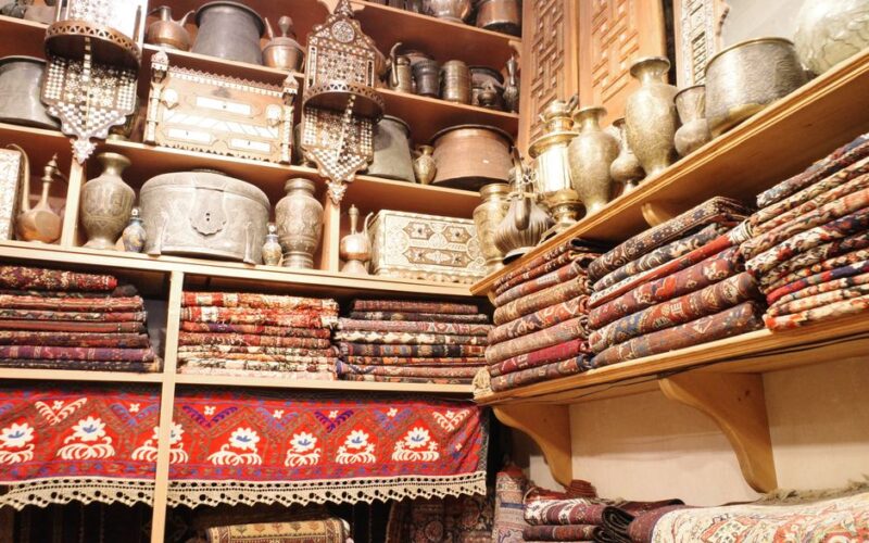 A vibrant scene showcasing folded Persian carpets and antique metalware in a traditional store setting.