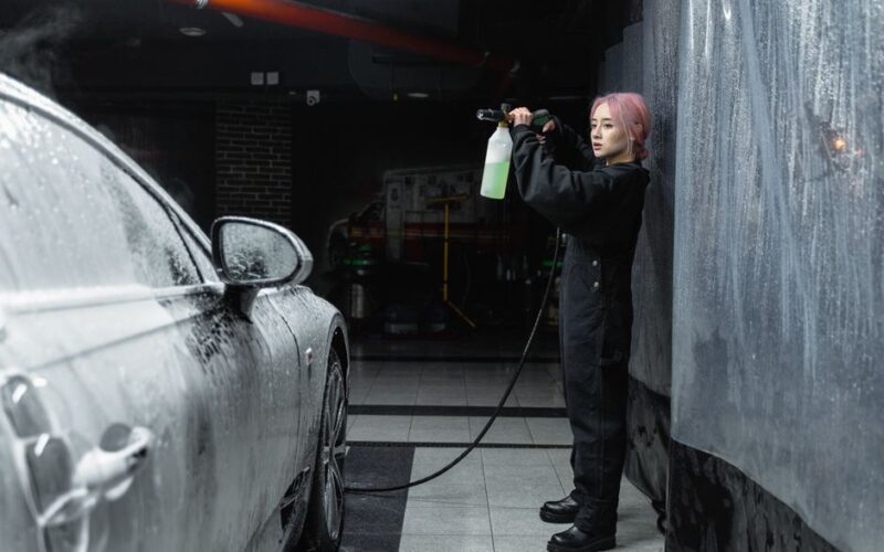 A young woman uses a power spray to clean a car with foam in an indoor garage.