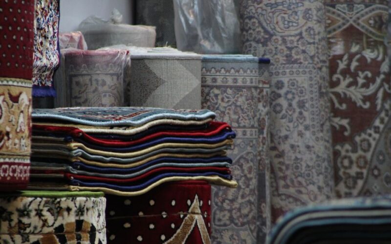 Vibrant folded and rolled rugs displayed in a traditional indoor bazaar setting.
