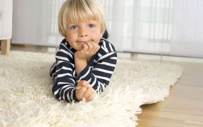 Adorable young boy with blonde hair laying on a white fluffy carpet indoors.