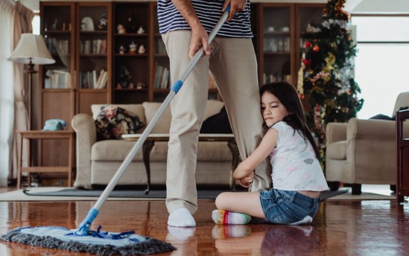 Father mopping the floor while daughter holds on, showing family bonding in a cozy living room.