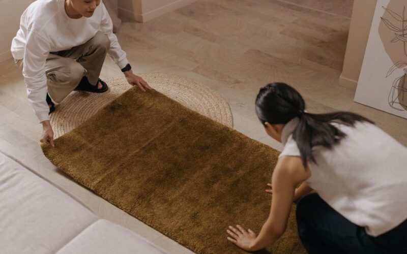 A man and woman collaboratively arranging a brown rug on a wooden floor inside a home.