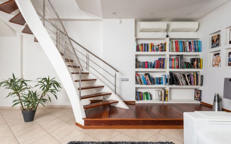 Assorted textbooks on shelves against contemporary staircase above ceramic floor with potted plant and carpet at home