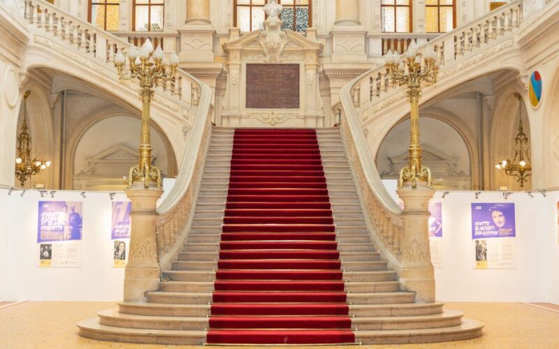 Elegant red carpeted staircase in a historic European interior, featuring ornate architecture and classic chandeliers.