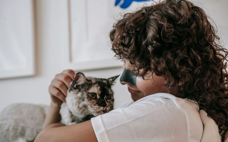 Side view of content young lady with dark curly hair and charcoal cleansing strip on nose smiling while hugging loyal cat at home