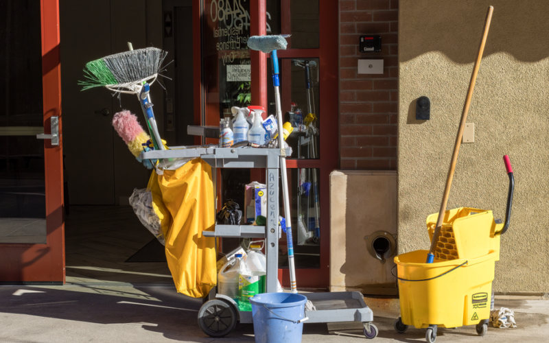 Cleaning supplies and tools outside a building entrance bathed in sunlight.