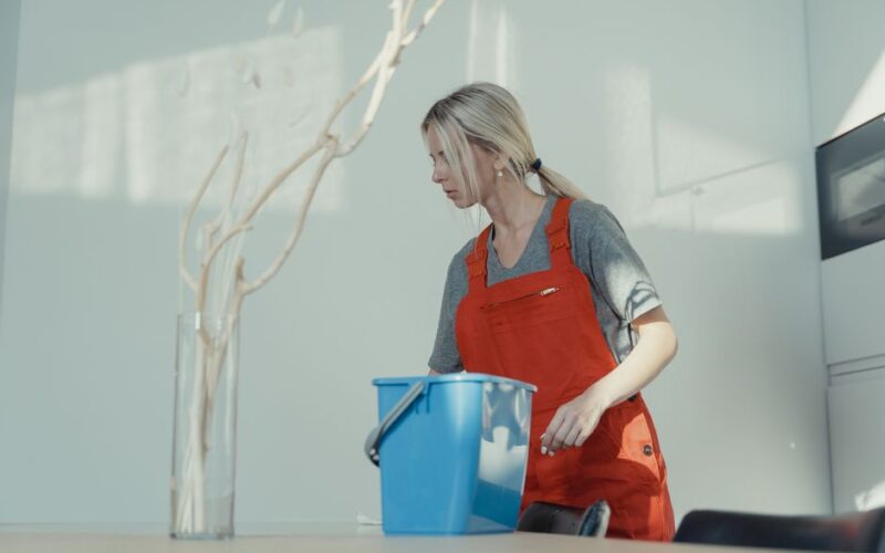 A woman in a red uniform cleans indoors with a blue bucket, ensuring a spotless environment.