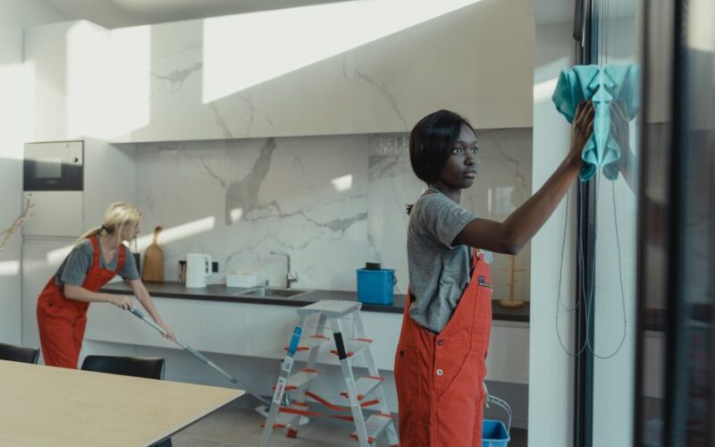 Two women cleaning a modern kitchen, wiping glass surfaces and mopping the floor.
