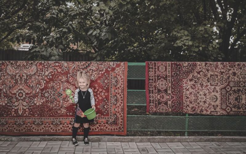 A young child smiling with a toy bucket in front of colorful rugs outdoors in Staromyshastovskaya, Russia.