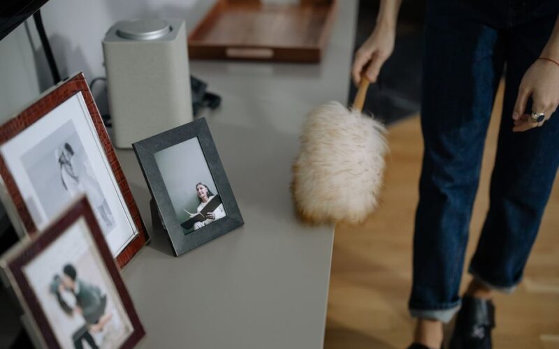 A woman using a feather duster to clean picture frames on shelves indoors.