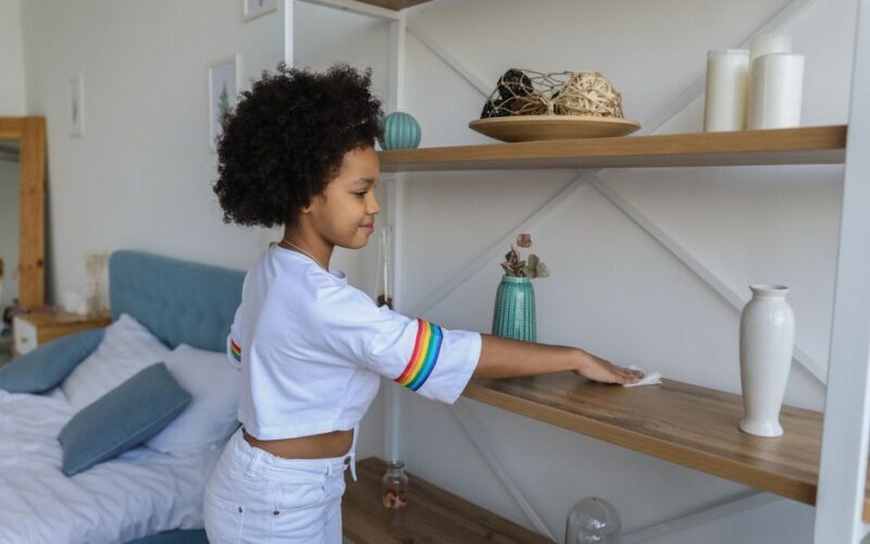 Charming African American girl wiping dust from wooden shelf of rack at home