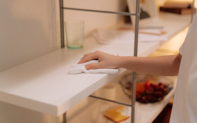 A person cleans a white shelf with a cloth, focusing on home maintenance.