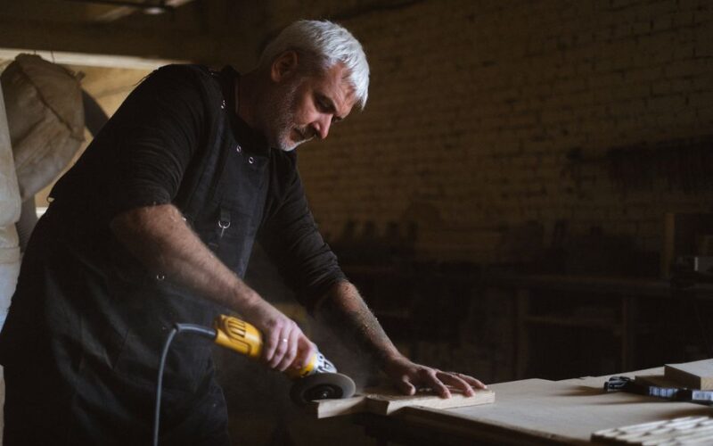 Side view of skilled male master in apron using electric angle grinder to polish wooden plank at table in professional carpentry