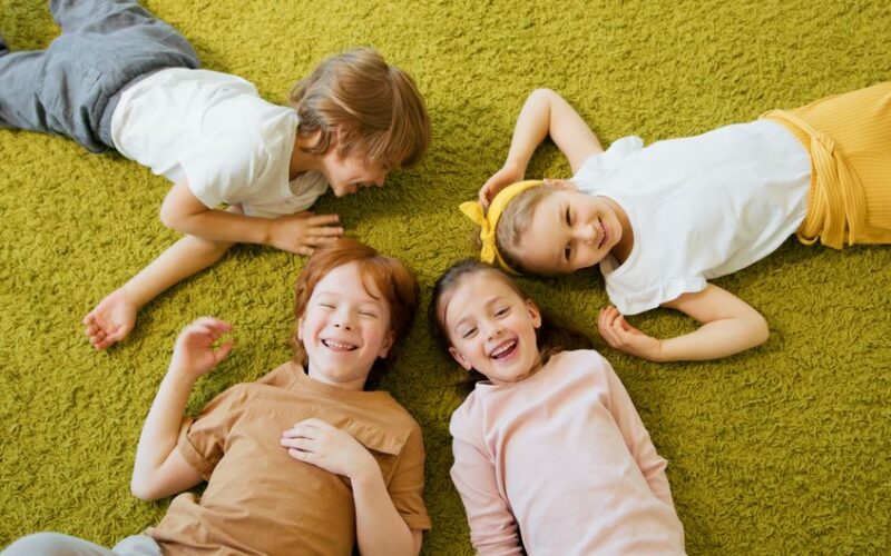 Four cheerful children lying together on a green carpet, enjoying their time indoors.