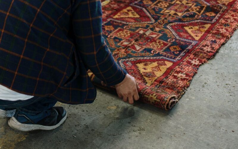 Close-up of an adult unrolling a detailed ornate carpet on a concrete floor.