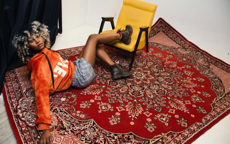 Stylish young woman in chic outfit posing in a studio on a decorative red rug next to a yellow chair.