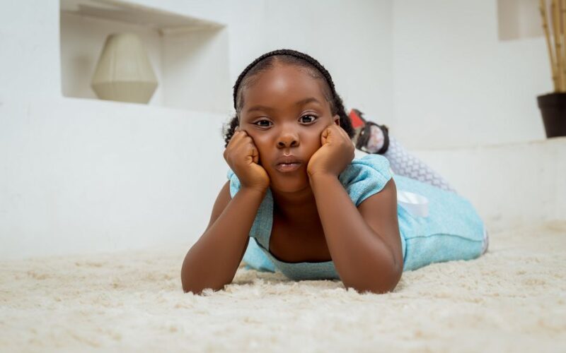 A young girl in a blue dress lying on a soft white carpet, indoors.