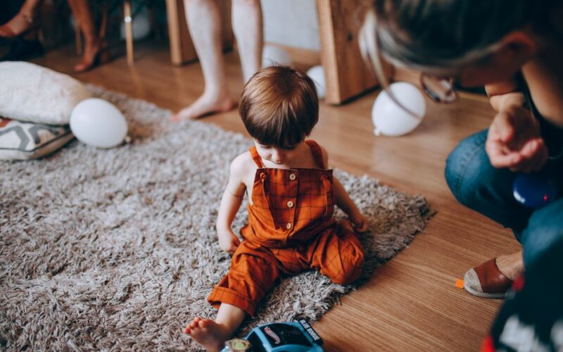 Child in orange overalls sits on a rug at an indoor birthday party with balloons around.
