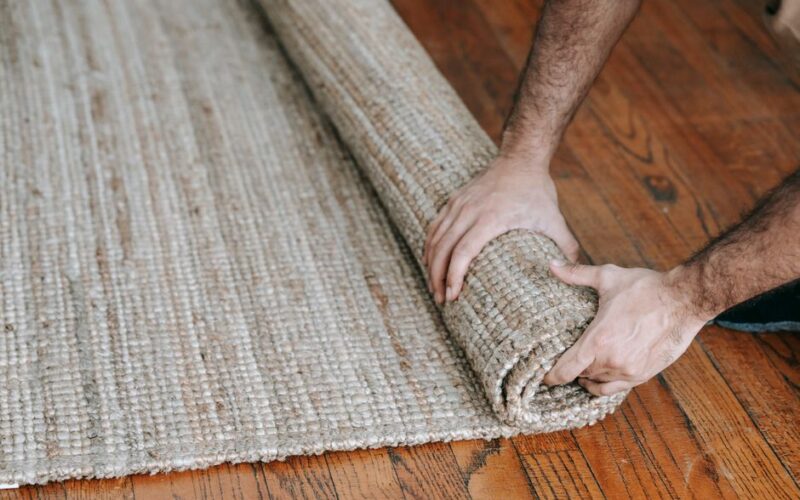 A man rolls up a textured woven rug on a wooden floor, indicating moving or cleaning.