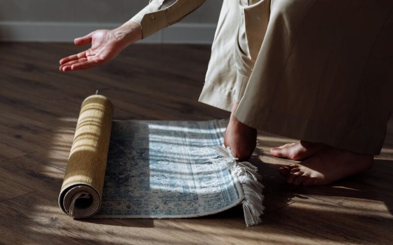 A person rolling out a prayer rug in a room with sunlight casting shadows on the floor.