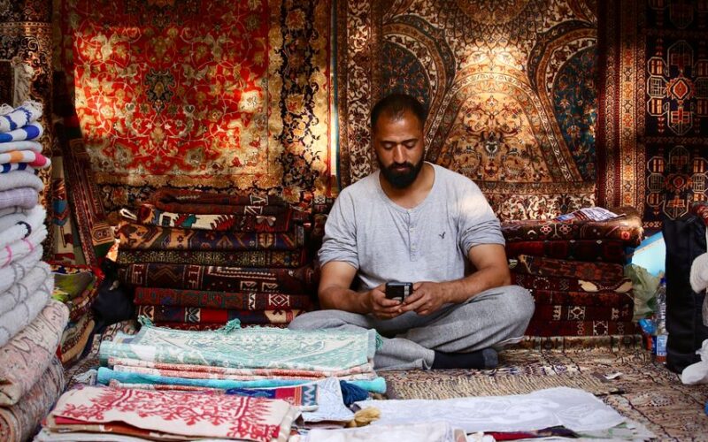 Middle-aged man in a carpet shop using phone amidst traditional textiles.