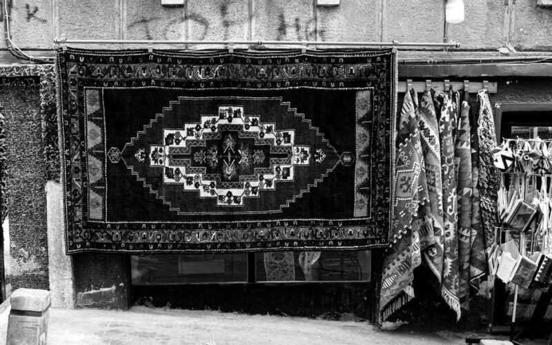 Black and white photo of traditional handwoven rugs displayed in an outdoor market setting.