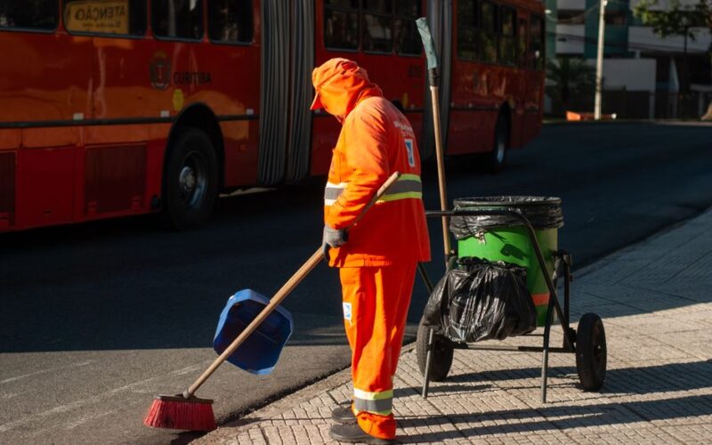Street cleaner in bright orange uniform sweeping sidewalk near public bus in city setting.