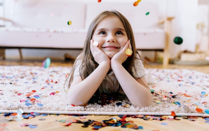Adorable child lying on carpet in a confetti-filled room, looking up with a joyful expression.