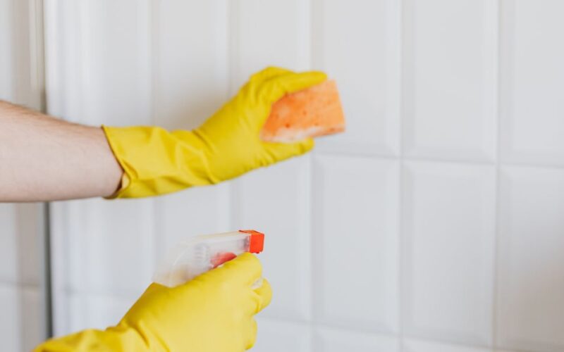 Crop unrecognizable woman in yellow rubber gloves cleaning white tiles with spray and soapy sponge