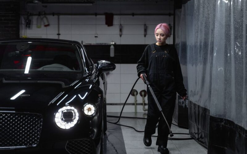 Woman cleaning luxury black car at car wash garage using hosepipe.