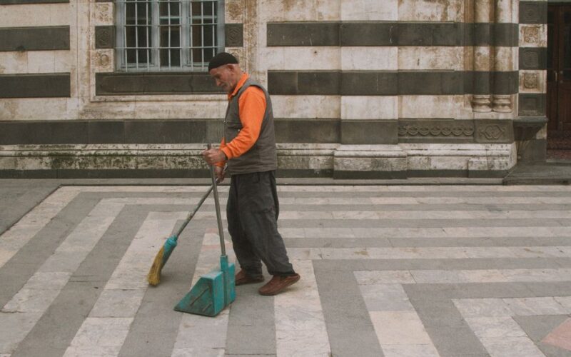 Elderly man sweeping courtyard in an urban setting with classic architecture.
