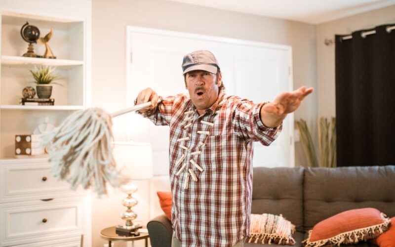 A man in a checkered shirt and cap entertains with a mop in a cozy living room.