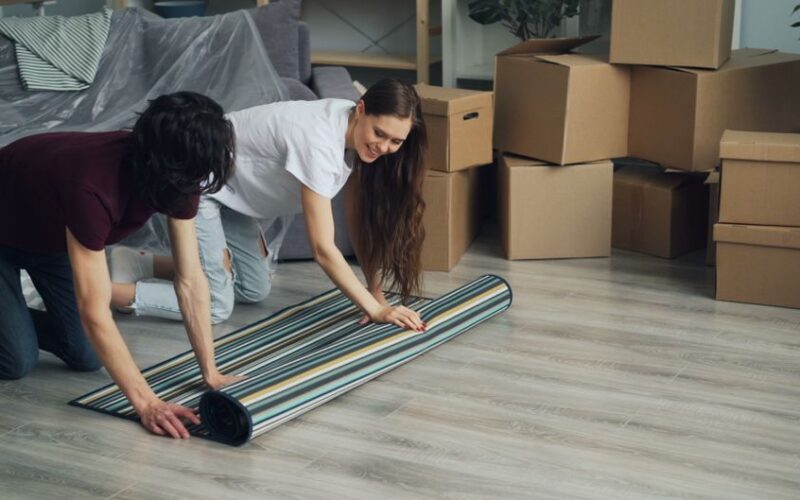 Couple smiling as they unroll carpet surrounded by moving boxes in their new home.